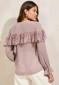 Dusty pink blouse with textured polka dots, lace ruffle trim, and puffed sleeves. Soft fabric, slight sheen, delicate details.