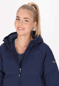 Námořnická modrá zateplená bunda se zipem vpředu, nastavitelnou kapucí a logem na rukávu. Nalezena přes pruhované tričko.