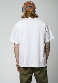 T-shirt blanc à manches courtes avec des manches raglan, texture lisse et coupe décontractée. Porté avec un pantalon vert olive et un bonnet tricoté multicolore.