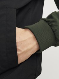 Crna jakna s zelenim rebrastim manžetama. Jakna ima bočnu džepicu, ističući glatku teksturu i pojednostavljeni dizajn.