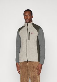 Modelo masculino vestindo um colete de fleece cinza e oliva com fecho zip, sobre uma camisa cinza de manga longa e calças de corduroy castanhas, de pé contra um fundo simples.
