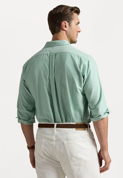 Gai zaļš pogājamais krekls ar sarullētām piedurknēm, aizmugures plecu dūriens detaļa, kombinēts ar baltām biksēm un brūnu jostu. Gluda auduma tekstūra.