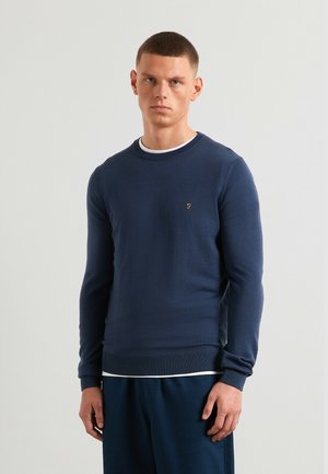 Pull en maille bleu marine avec un col rond, des poignets et un bas côtelés. Présente un logo discret sur la poitrine gauche, porté par-dessus une chemise blanche.