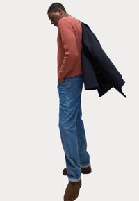 Suéter acanalado de color óxido, vaqueros de mezclilla azul y zapatos de ante marrón. Un abrigo azul marino colgado del hombro añade contraste al conjunto.