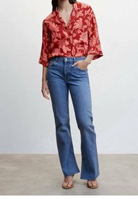 Camicia rossa a maniche corte con motivo floreale, abbinata a jeans a vita alta a zampa d'elefante blu. Indossata con sandali con cinturino beige.