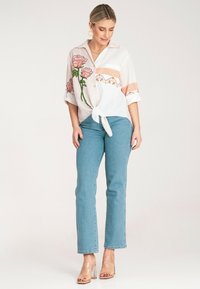 Biała bluzka z różowym haftem kwiatowym, w paski, krótki rękaw, związana w talii. Jasnoniebieskie proste dżinsy i przezroczyste sandały na obcasie.