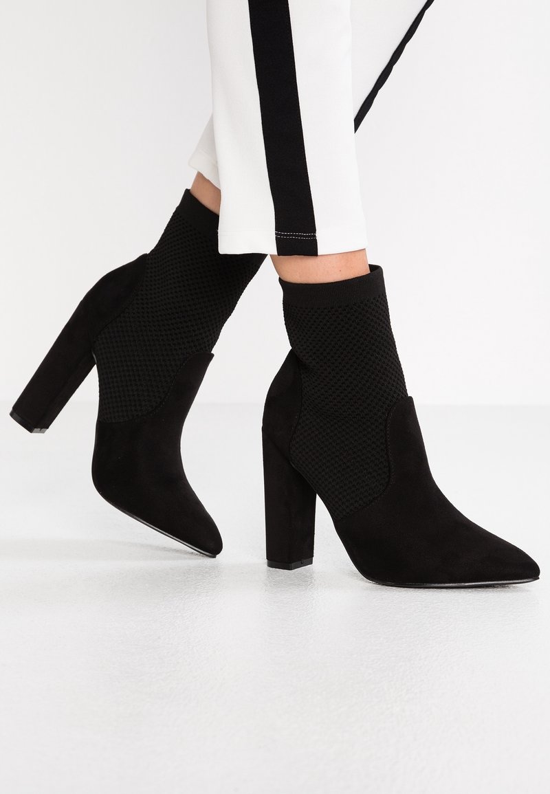 Bottines à talon bloc en tricot noir et suède portées avec un pantalon blanc présentant une bande latérale noire sur un fond de sol blanc.