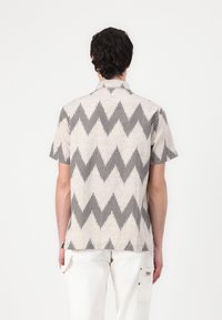 Camicetta a maniche corte di un beige chiaro con motivo zigzag nero, realizzata in tessuto testurizzato, con vestibilità rilassata e colletto.