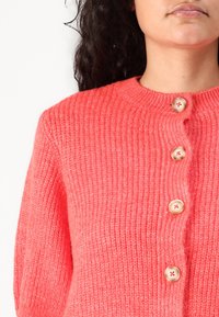 Korálově růžový pletený kardigan s kulatým výstřihem a čtyřmi krémovými knoflíky vpředu s žebrovanou strukturou po celém kardiganu.