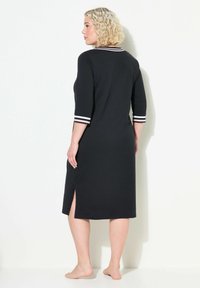 Černé šaty po kolena s tříčtvrtečními rukávy, které mají bílé pruhy na manžetách a boční rozparek. Hladká tkanina a volný střih.