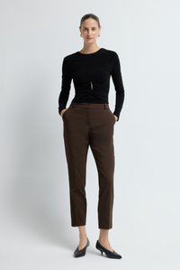 Vrouw staat met handen in de zakken, gekleed in een zwarte lange mouwen ruche top, bruine op maat gemaakte broek en zwarte lage hakken voor een witte achtergrond.