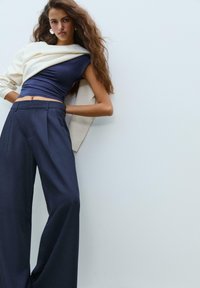 Top blu navy attillato e pantaloni a gamba larga in tessuto scuro. Un maglione oversized color crema drappeggiato sulle spalle. Texture liscia e linee pulite.