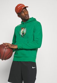 Zelená mikina s kapucňou a logom Boston Celtics, zmes bavlny, predsadená vrecko a nastaviteľná šnúrka. Čierne športové šortky s bielym logom.