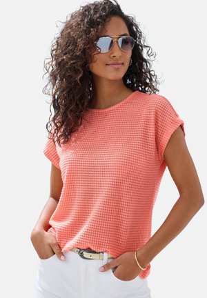 Frau mit lockigem Haar trägt verspiegelte Sonnenbrille, korallenfarbenes, strukturiertes Kurzarmoberteil, weiße Hose und einen leichten Gürtel, Hände in den Taschen.