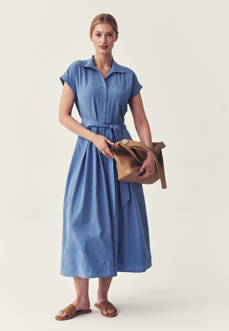 Femme en robe midi bleue boutonnée avec taille nouée, tenant un sac à main beige et portant des sandales marron sur un fond clair uni.