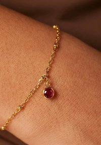 Gouden schakelarmband met een ronde, facette rode edelsteen hanger, rustend op een pols. De ketting heeft kleine gestructureerde schakels.