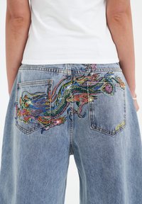 Light-wash denim jeans featuring a colorful, beaded dragon design on the back pockets and a relaxed fit with an unfinished hem.