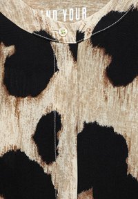 Tessuto con stampa leopardata in beige e nero. Presenta un collo rotondo e una chiusura con bottone vicino al colletto. Texture liscia con un design moderno.