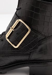 Bota de cuero negra con textura de cocodrilo, que presenta una hebilla cuadrada dorada y costuras detalladas en la correa del tobillo.