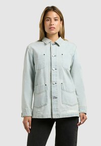 Femeie cu păr drept, castaniu deschis, purtând o jachetă din denim deschis cu patru buzunare frontale și pantaloni negri, pe un fundal simplu.