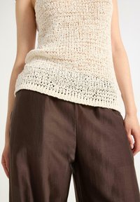 Sweter w kolorze ecru z luźnym splotem; zestawiony z brązowymi, błyszczącymi spodniami typu wide-leg. Skoncentruj się na fakturze i kontrastach kolorystycznych między elementami odzieży.