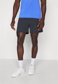 Mand iført blå træningsskjorte, sorte shorts med lille orange logo, hvide sokker og hvide sneakers, der står mod en hvid baggrund.