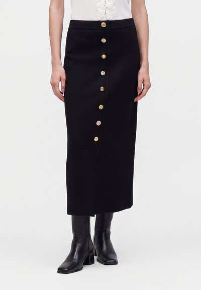 Woman wearing a black buttoned midi skirt with gold buttons, black heeled boots, and a white top with ruffle detail.
