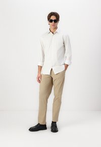 Camisa de botão branca e bege com riscas, calças bege e sapatos pretos. Modelo a usar óculos de sol, em pé contra um fundo branco liso.