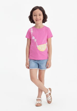 Χαμογελαστή κοπέλα με κοντά σκούρα μαλλιά, φορώντας ροζ μπλουζάκι με σχέδιο λεμονιού και λουλουδιών, τζιν σορτς και λευκά σανδάλια, όρθια με σταυρωμένα πόδια.