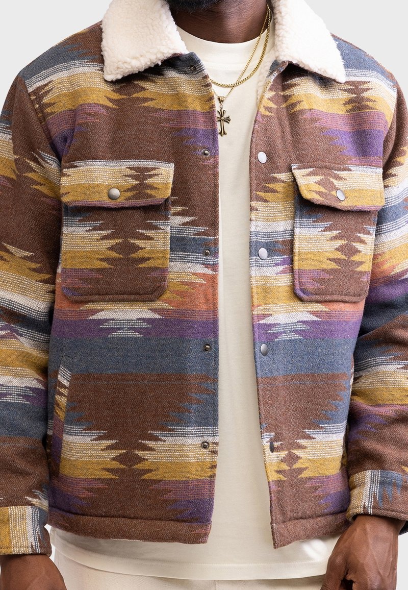 Veste marron à motifs multicolores avec un col en shearling, dotée de deux poches poitrine boutonnées et de fermetures à pression. Tissu texturé.