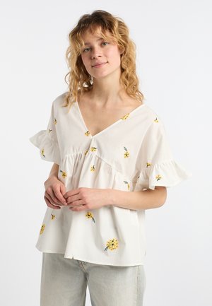 Junge Frau mit lockigem blondem Haar, die eine weiße Bluse mit gelber Blumenstickerei und Rüschen an den kurzen Ärmeln trägt, kombiniert mit hellen Hosen.