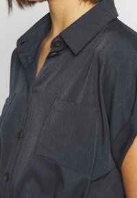 Chemise bleu foncé à manches courtes avec col, deux poches poitrine et boutons en bois. Le tissu présente un léger éclat et une texture douce.