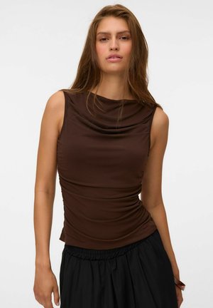 Jeune femme aux longs cheveux portant un haut marron sans manches et un pantalon noir ample, debout devant un fond clair uni.