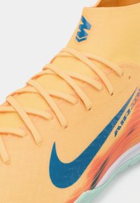 Detailní záběr na oranžové tenisky Nike Air Zoom s modrým znakem swoosh a odpovídajícími oranžovými tkaničkami na světlém pozadí.