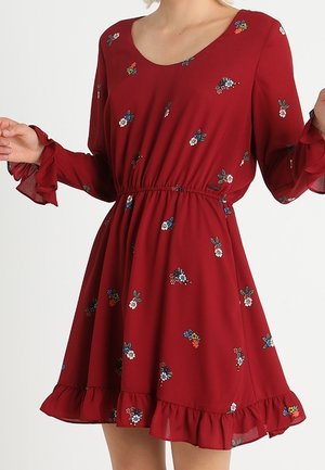 Rød knælængde kjole med lange ærmer, elastisk talje, flæsekant og små flerfarvede blomsterprint spredt over stoffet.