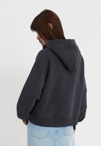 Mørkegrå hettegenser med stor hette, senkede skuldre og ribbestrikket kant, kombinert med lyse denimjeans. Enkel, uformell design.
