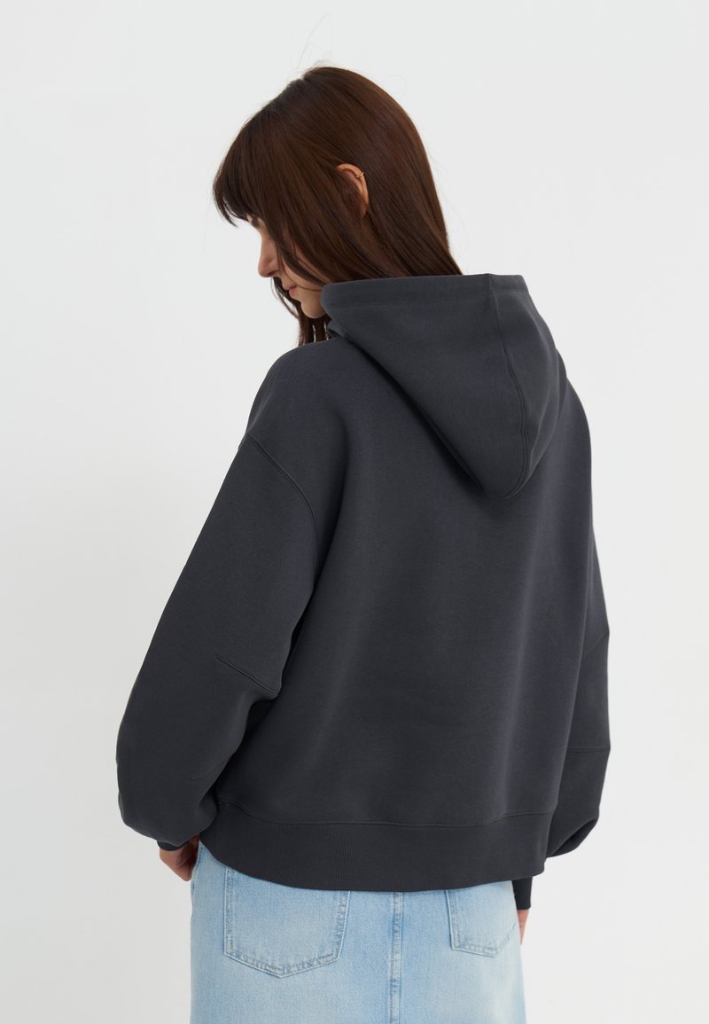 Ciemnoszara bluza z dużym kapturem, opadającymi ramionami i ściągaczem na dole, połączona z jasnymi dżinsami. Prosty, casualowy design.