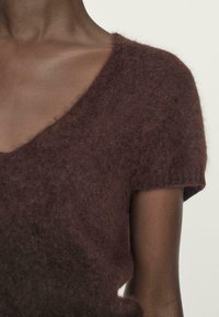 Maglione corto a manica corta marrone con scollatura a V profonda, caratterizzato da una texture morbida e pelosa. Dettaglio a coste sui polsini delle maniche.