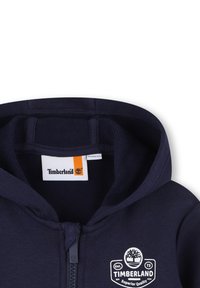 Felpa con zip di colore blu navy, realizzata con un tessuto morbido, caratterizzata da un patch con logo Timberland, un'etichetta arancione e un design classico del cappuccio.