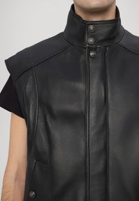 Μαύρο δερμάτινο γιλέκο με ψηλό γιακά, κουμπιά με κούμπωμα και κοντά μανίκια. Διατίθεται με λεία υφή και πλευρικές τσέπες.