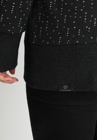 Primo piano di una manica e di un orlo di un maglione nero con piccoli motivi a cuore pastello e un polsino a costine, indossato con pantaloni neri.