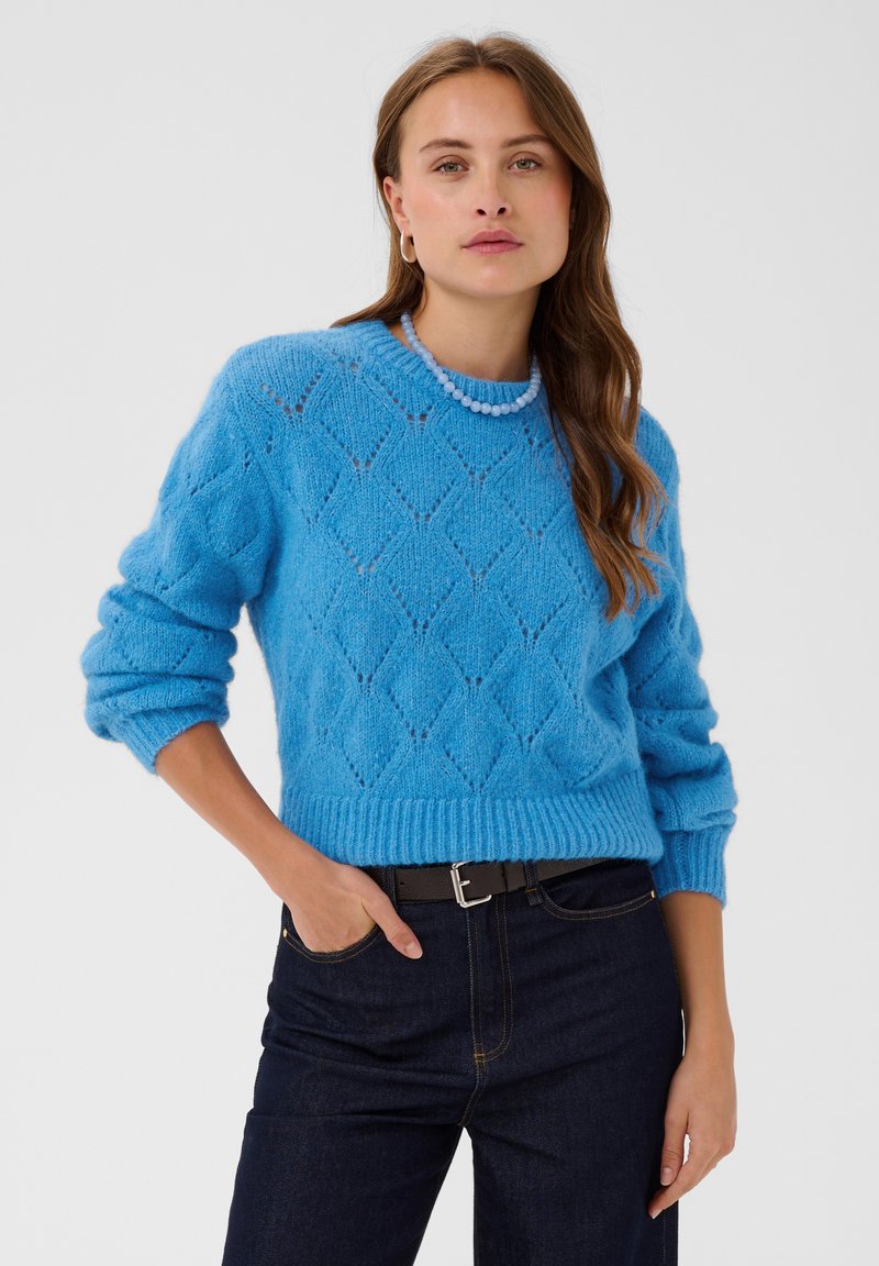 Femme aux longs cheveux bruns debout portant un pull bleu texturé, un jean foncé, un collier de perles bleues et des boucles d'oreilles dorées en anneau sur un fond uni.