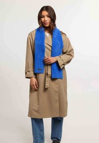 Trench coat beige con vita a cintura, abbinato a una sciarpa pelosa blu brillante. Jeans in denim completano l'outfit.