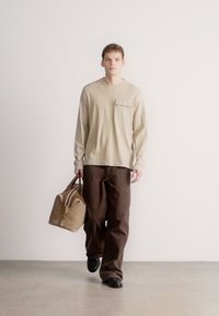 Béžová košeľa s dlhým rukávom a náprsným vreckom, v kombinácii s voľnými hnedými nohavicami. Model drží hnedo-béžovú tašku s hnedými detailmi. Jednofarebné pozadie.