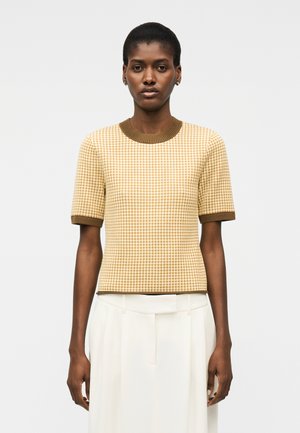 Γυναίκα με σκουρόχρωμο δέρμα και κοντά μαλλιά, φοράει μπεζ και καφέ καρό πουλόβερ με κοντά μανίκια και λευκή πλισέ φούστα, στέκεται με το σώμα στραμμένο προς τα εμπρός.