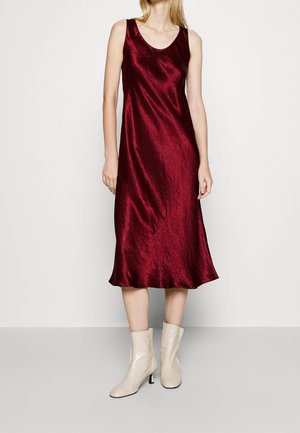 Woman wearing sleeveless, knee-length, deep red satin dress with scoop neck and off-white ankle boots, standing against plain background.