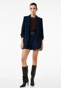Marineblazer over een bruine top, gecombineerd met marineblauwe shorts en hoge zwarte leren laarzen. De blazer heeft een gestructureerd ontwerp en opgerolde mouwen.