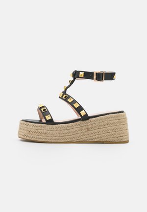 Sorte platform sandaler med en vævet jutesål, der har gyldne pyramide-nitter på stropperne og en spænde-lukning ved anklen.