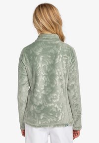 Chaqueta de forro polar en verde pálido con un patrón floral texturizado, que presenta un cuello alzado y mangas largas.