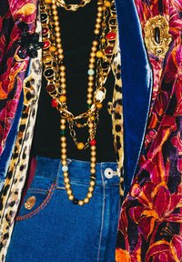 Blazer coloré et à motifs, orné de colliers à chaîne dorée superposés sur un haut noir. Jean en denim bleu taille haute avec un détail de broderie de rose.